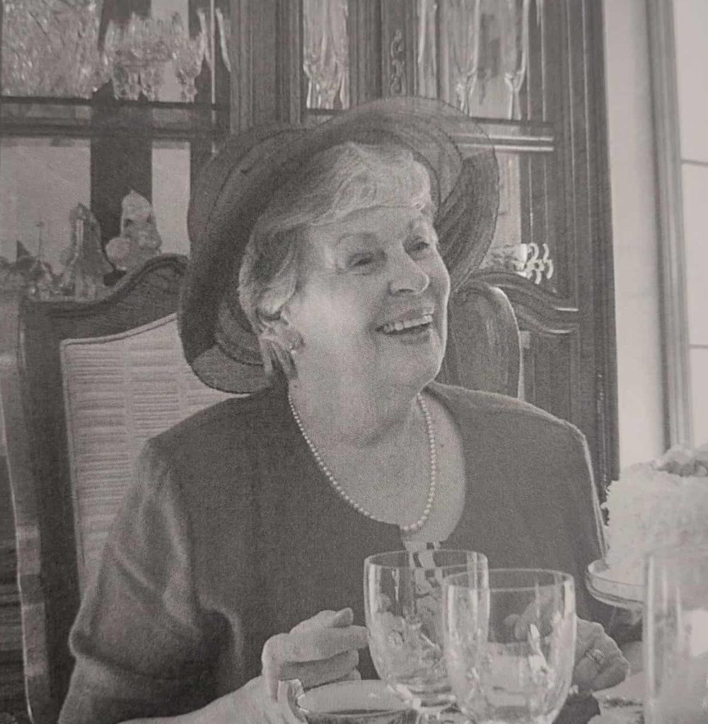 Black and white image of a woman sitting at a table. On the table are crystal glasses. The woman smiles and wears a big hat and a pearl necklace