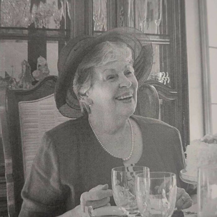 Dawn Anderson MacFarlane, Date Unknown, According to Me by Dawn Anderson MacFarlane Black and white image of Dawn MacFarlane sitting at a table. On the table are crystal glasses. The woman smiles and wears a big hat and a pearl necklace