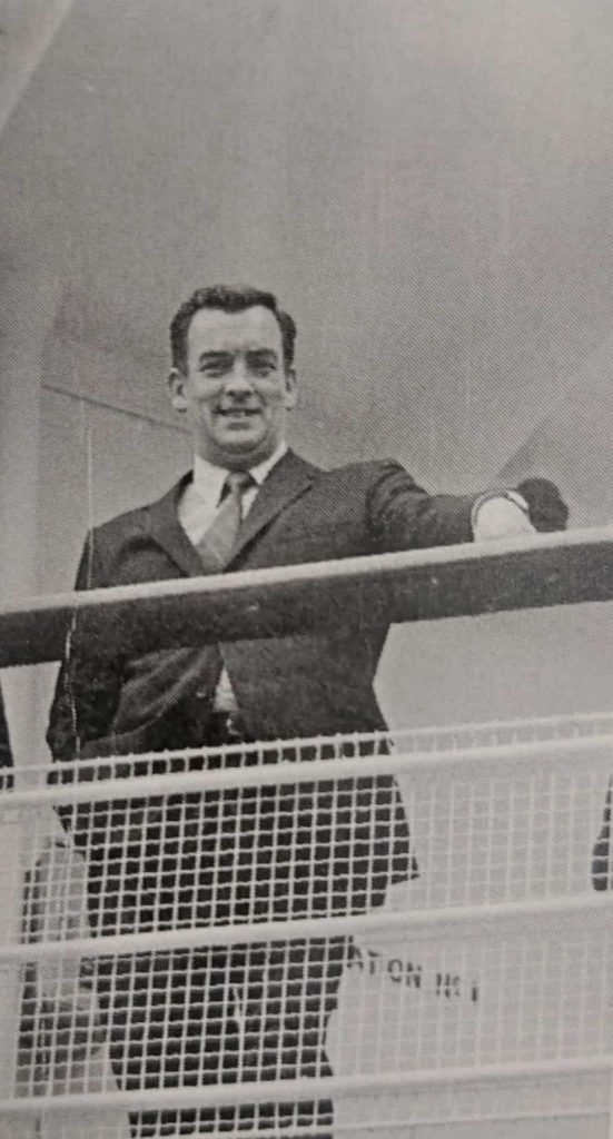 Black and white image of a man standing on a boat deck. His hand rests on the rail