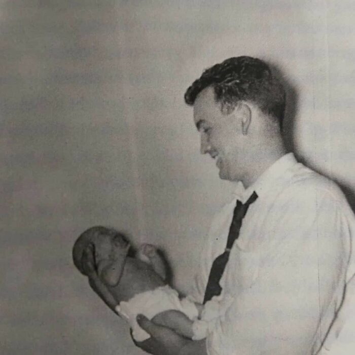 Mine! 1957, According to Me, Dawn Anderson MacFarlane Black and white image of a man holding a newborn baby. He smiles at the baby