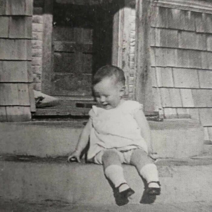 On Steps of Anderson Home at 14 Months Old, 1931, According to Me by Dawn Anderson MacFarlane Black and white image of a toddler sitting on the front steps of a house. The toddler smiles while looking down and wears a white dress with white socks and black shoes