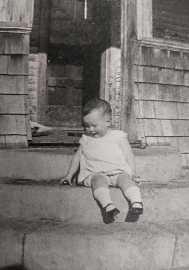 Black and white image of a toddler sitting on the front steps of a house. The toddler smiles while looking down and wears a white dress with white socks and black shoes