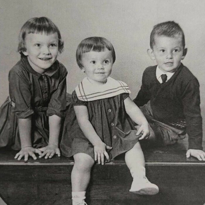 Portrait of Children 2, 4, 6, c1963, According to Me by Dawn Anderson MacFarlane Black and white image of three children sitting on a bench. The child on the left wears a dress and leans onto the bench. Her hair is short and her hands are splayed as she leans forward. THe child in the middle also wears a dress. The child on the right leans on one hand while sitting. He is wearing a sweater and a tie