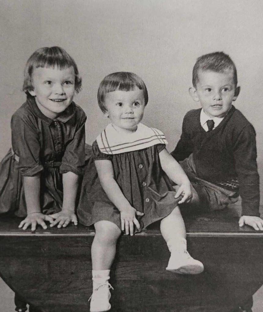 Black and white image of three children sitting on a bench. The child on the left wears a dress and leans onto the bench. Her hair is short and her hands are splayed as she leans forward. THe child in the middle also wears a dress. The child on the right leans on one hand while sitting. He is wearing a sweater and a tie