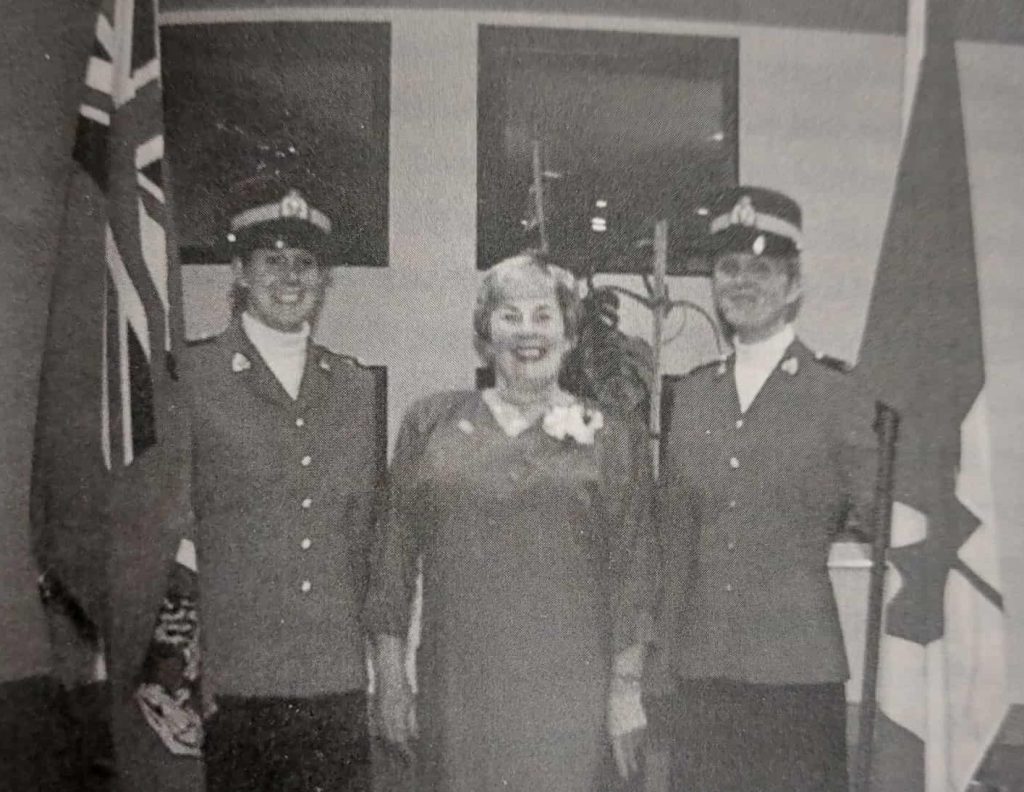 black and white image of three women standing together. The women on the right and on the left are in uniform and hold a British flag on the left and a Canadian flag on the right. In the center of the image Dawn MacFarlane stands with a huge smile
