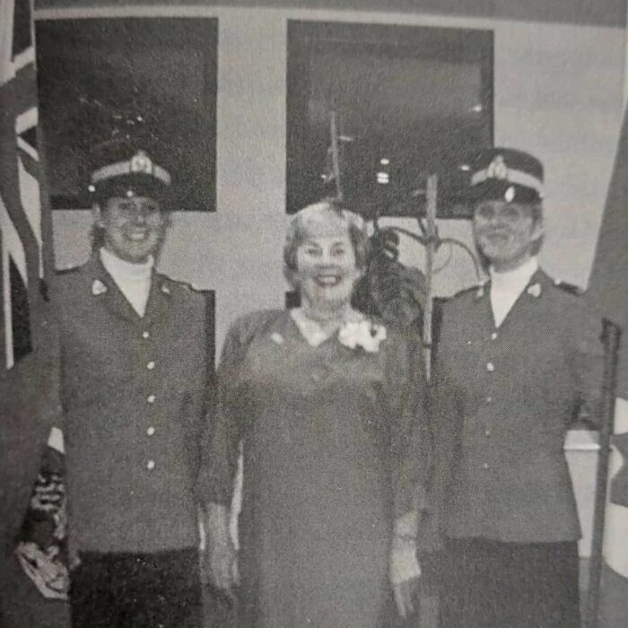 Swearing In Ceremony Deputy Mayor, 1981, According to Me by Dawn Anderson MacFarlane black and white image of three women standing together. The women on the right and on the left are in uniform and hold a British flag on the left and a Canadian flag on the right. In the center of the image Dawn MacFarlane stands with a huge smile