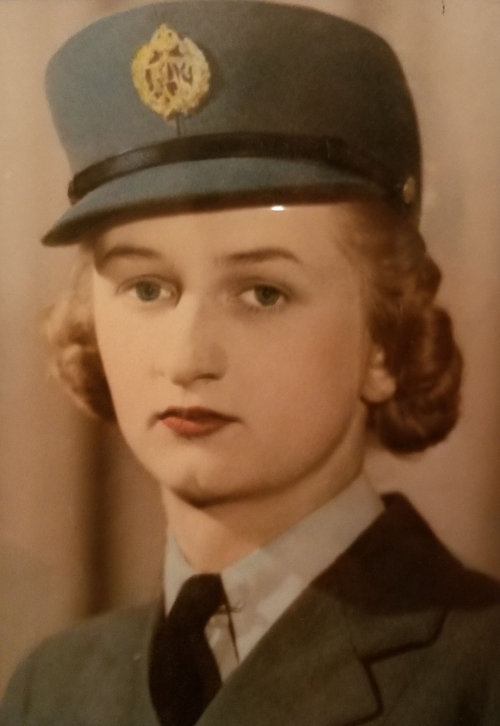 Colour image of Beatrice Gunter. She looks past the camera and wears a shirt, tie, and jacket. Her hair is pinned up and she wears a blue hat