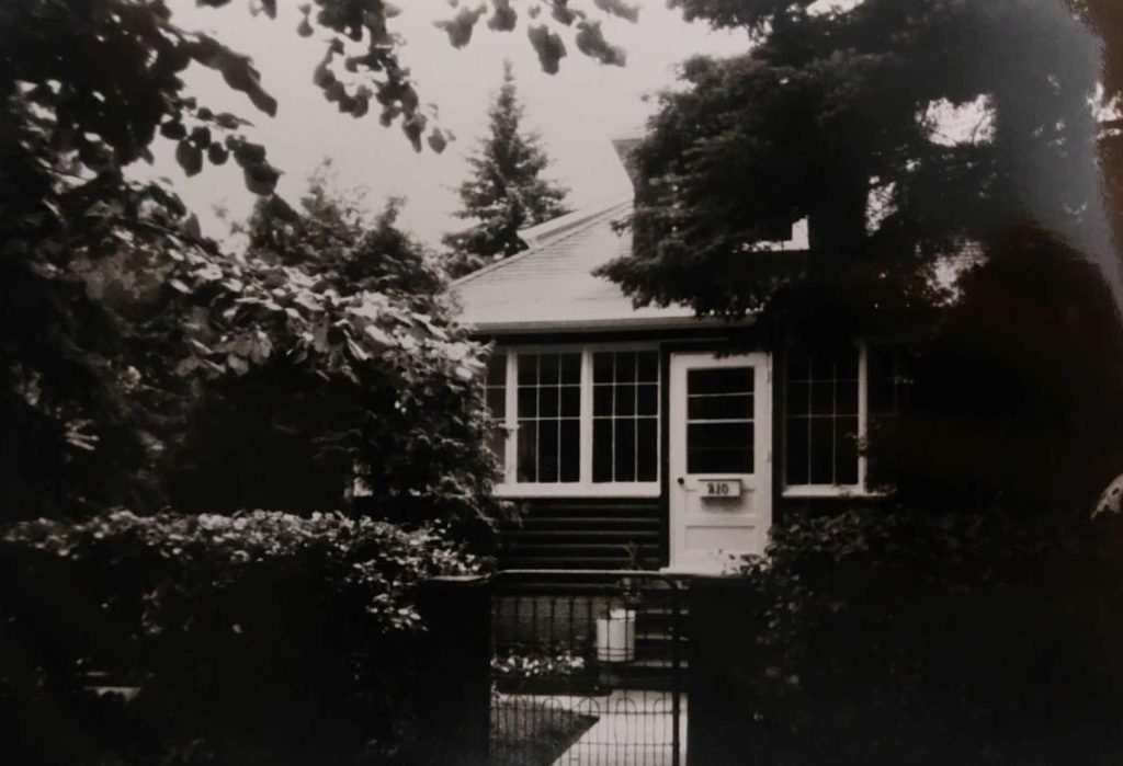 A black and white image of a house surrounded by trees. The house has a sunroom with windows and a white door