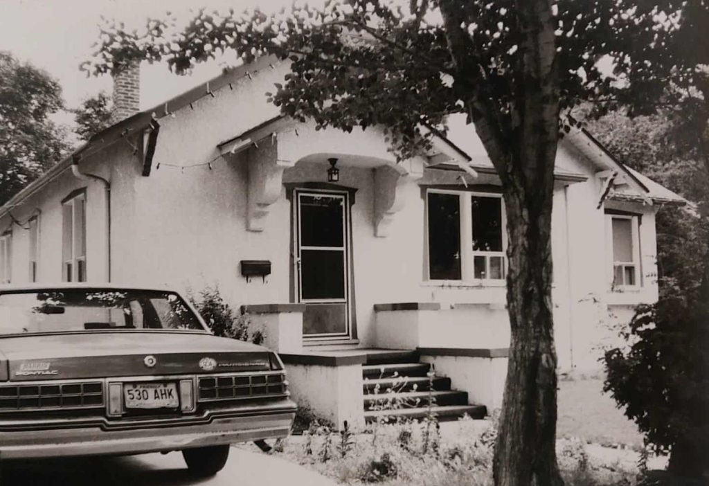 Black and white photo of a white house. Theres a car parked in the driveway and a tree overhanging the yard
