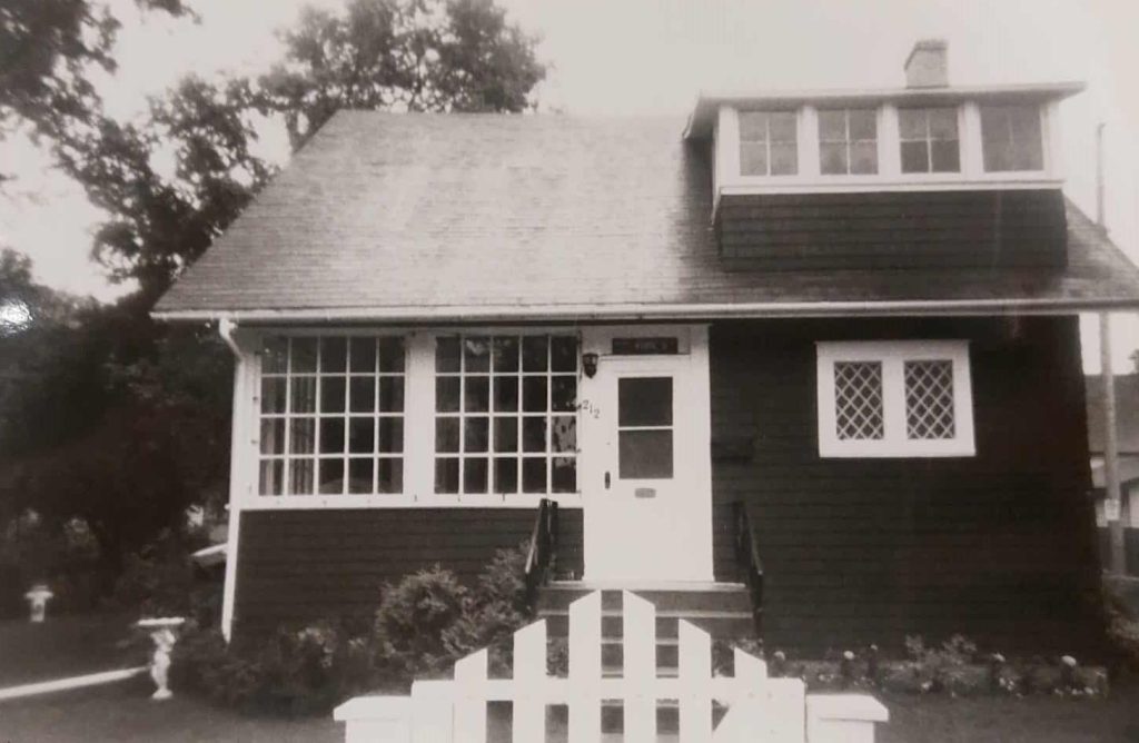 A black and white image of a house with white trim. There is a white picket gate at the end of the walkway up to the house