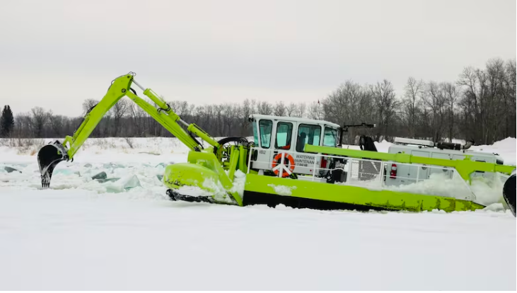 Colour image of an ice breaking machine. The machine is on the river working to break up the ice. The machine is bright neon green and has a cabin similar to a boat with an extended arm attachment similar to an excavator