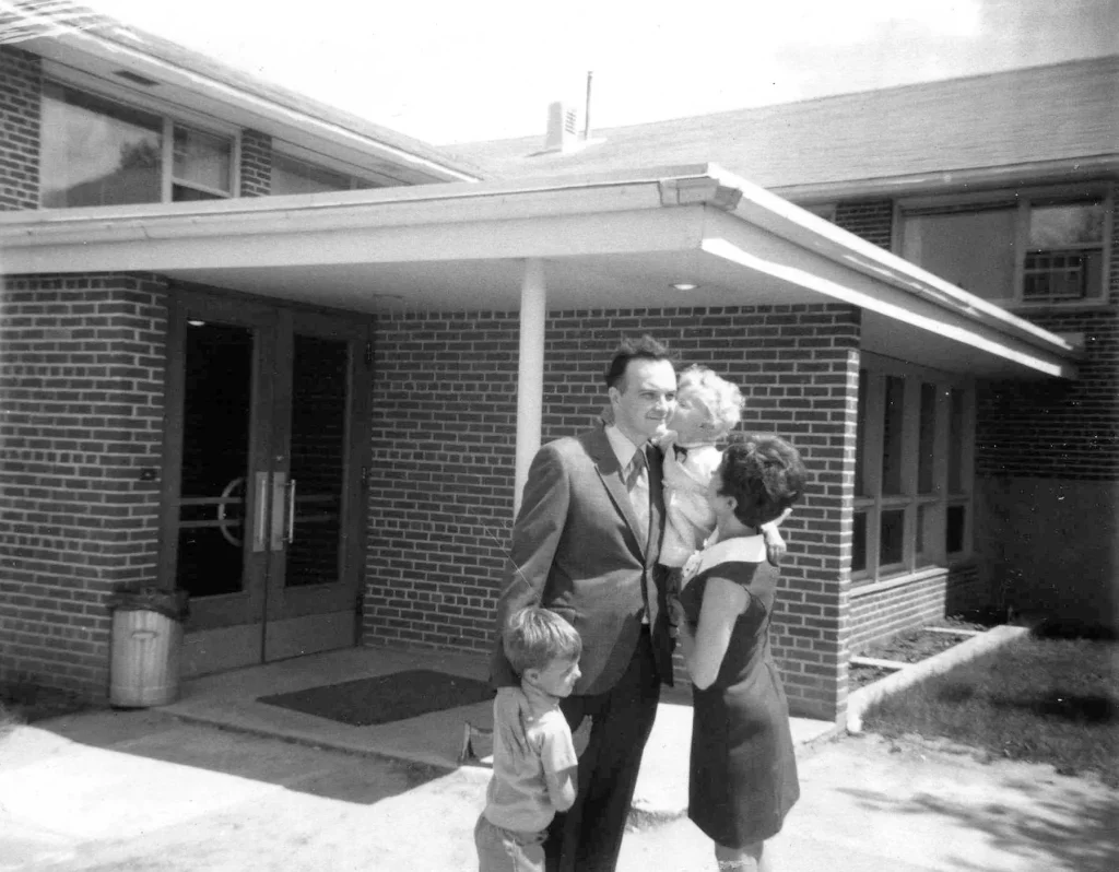 A black and white photo of a husband and wife with their two young children embracing just outside a brick building