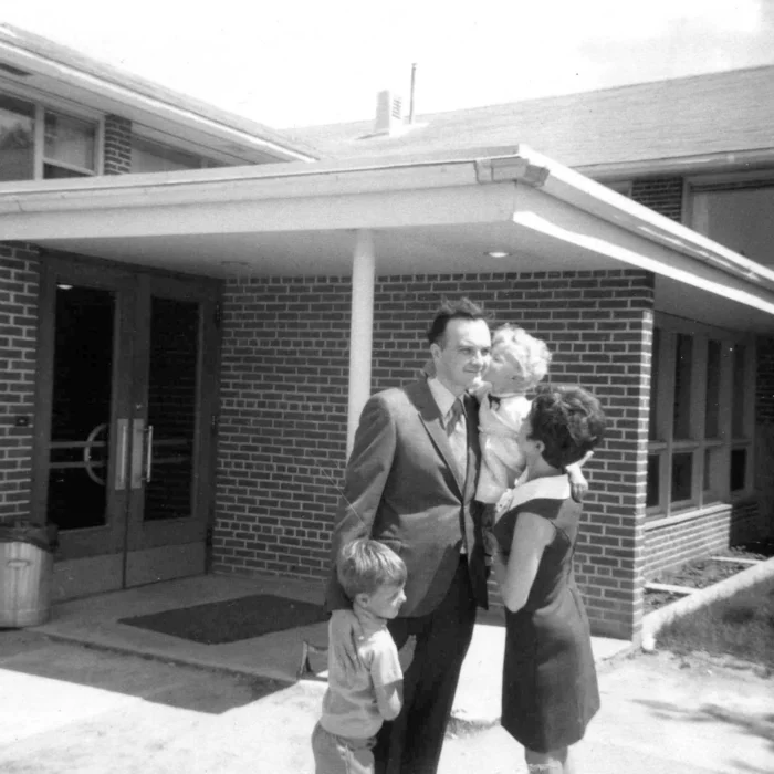 Howard Pawley and Family, 1969, Winnipeg Free Press A black and white photo of a husband and wife with their two young children embracing just outside a brick building