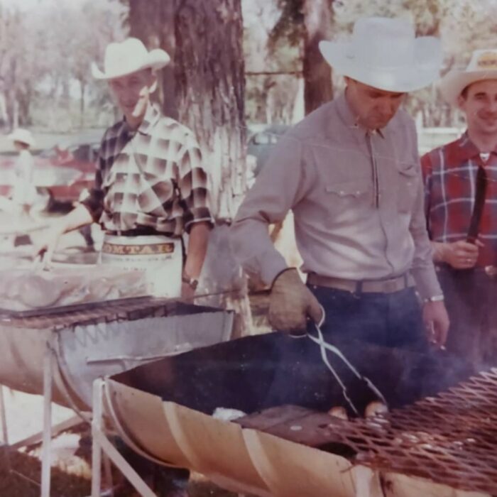 Colour image of a man working at a grill. There is a man in the background and to the right who are both wearing plaid. All the men are wearing white cowboy hats