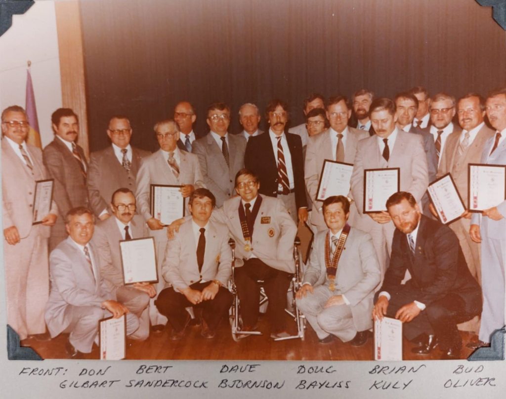 A colour image of a group of men sitting and standing in a large group. Some of them are holding certificates