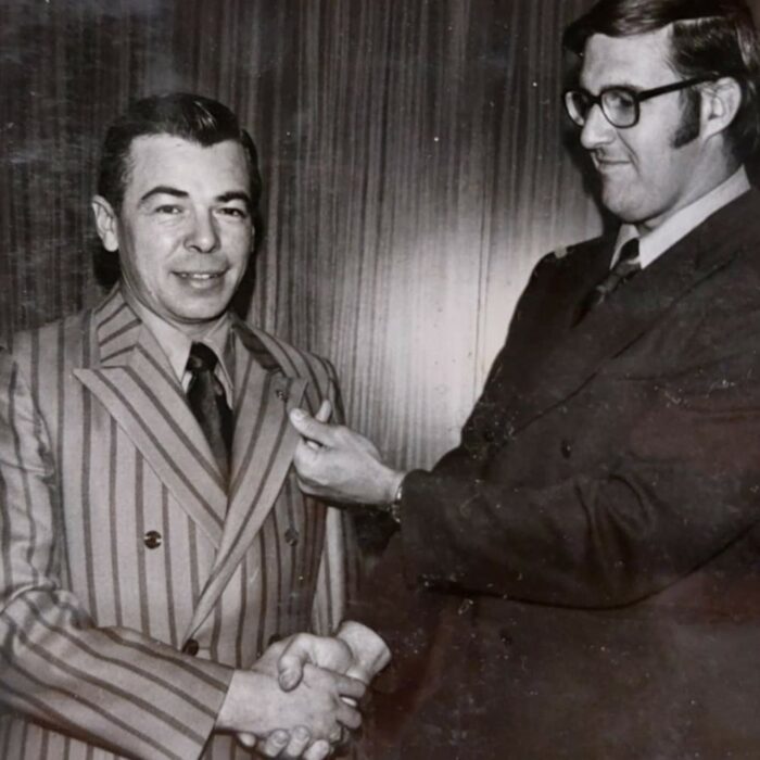 Black and white image of two men standing together. The man on the left has a striped suit on and the man on the right wears glasses and a black suit