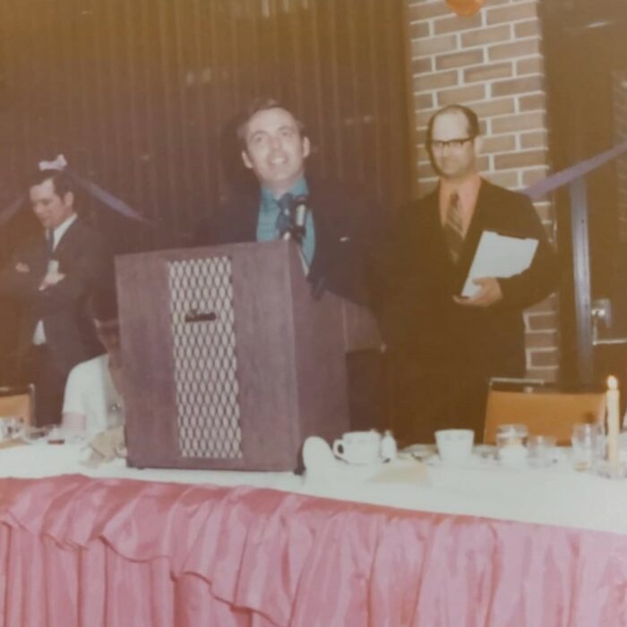 Colour image of three men. One stands at a podium, the other two are in the background watching