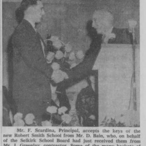 Mr. F Scardina Accepts Keys from Mr. D Bain for Robert Smith School Opening, 1959, Selkirk Enterprise Black and white image of two men shaking hands. One stands behind a podium. there are flowers in the background