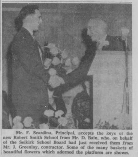 Black and white image of two men shaking hands. One stands behind a podium. there are flowers in the background