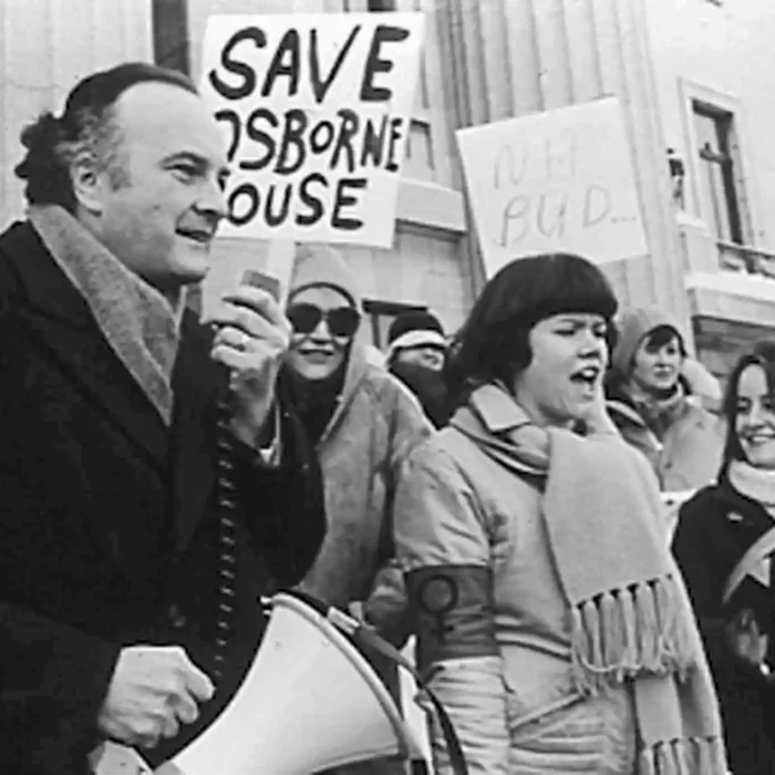 Pawley Addresses Protesters Calling for Longterm Funidng for the Osborne House Women's Centre, 1979, Winnipeg Free Press A black and white photo of Howard Pawley on the left hand side. He holds a megaphone and is about to speak into the receiver. Beside him are women dressed in jackets, scarves, and hats. One holds a sign which says Save Osborne House