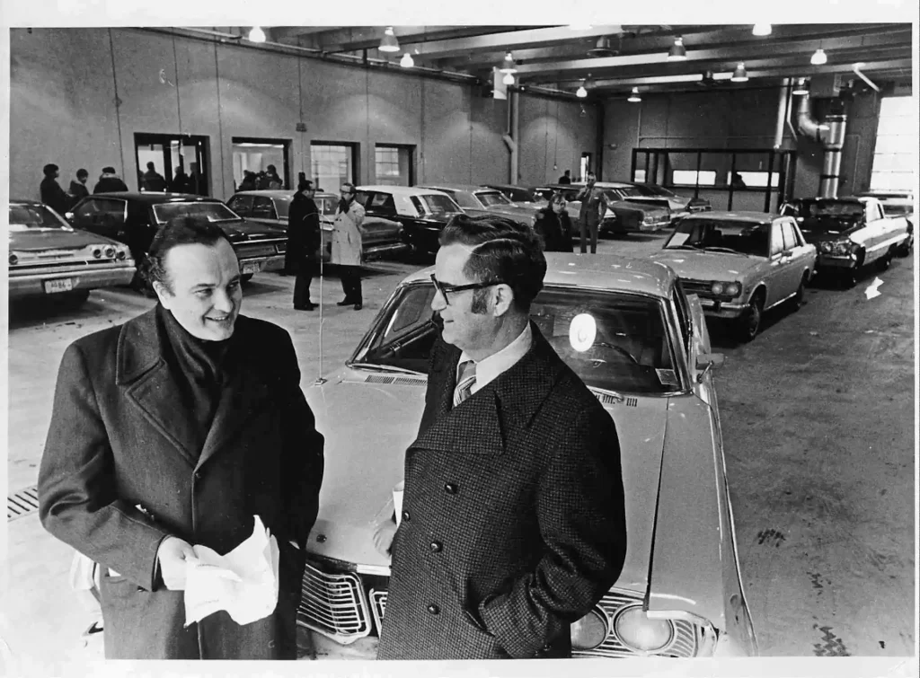 A black and white photo of Howard Pawley and another man standing inside a car garage. They are dressed in buttoned jackets