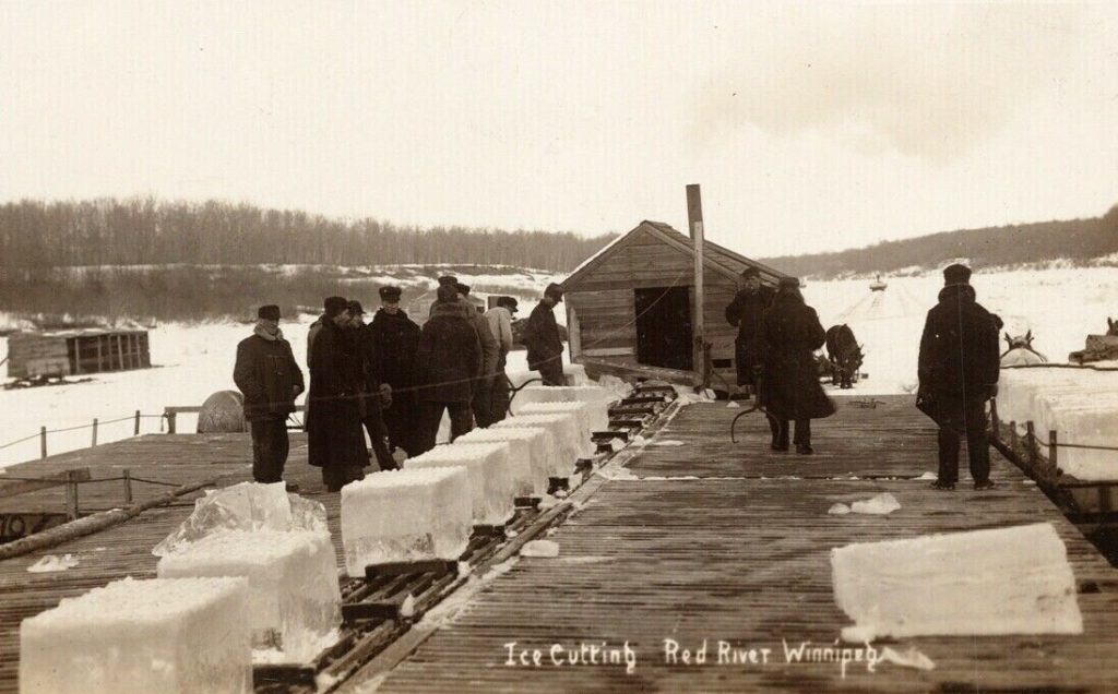 A sepia-coloured image of men standing next to many large blocks of ice on a conveyor. At the end of the conveyor is a small hut