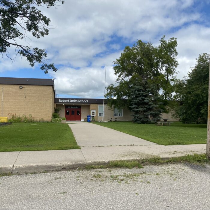 Robert Smith School, 2024, Selkirk Museum Collection Colour image of Robert Smith School. There are trees beside the entrance walkway and the front doors are a dark burgundy red. The school is brown brick and the sky is blue