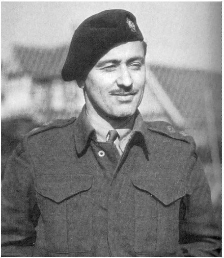 Black and white photo of a soldier in uniform. He wears a black hat, a moustache, and a tie. He looks to the right and smiles