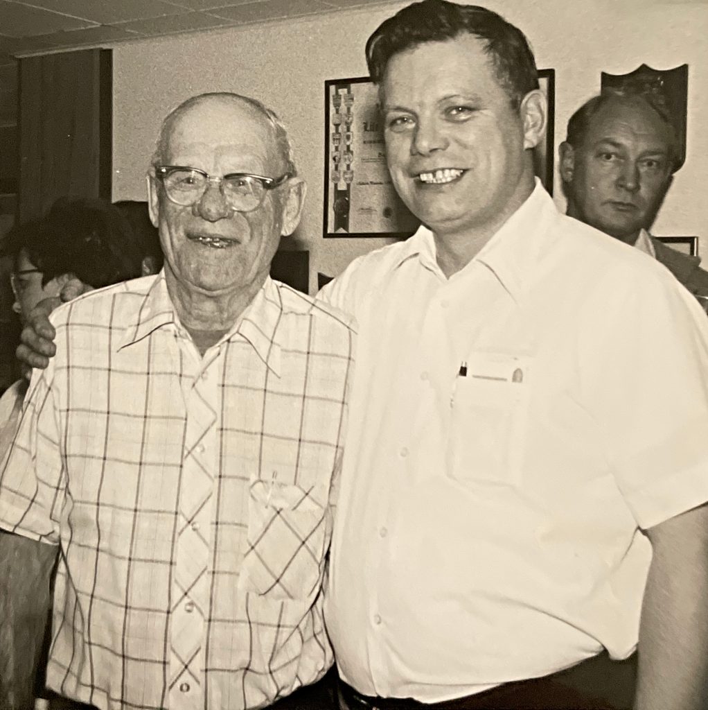 Black and white image of two men standing next to eachother. The younger man has his arm around the older man. Both men smile