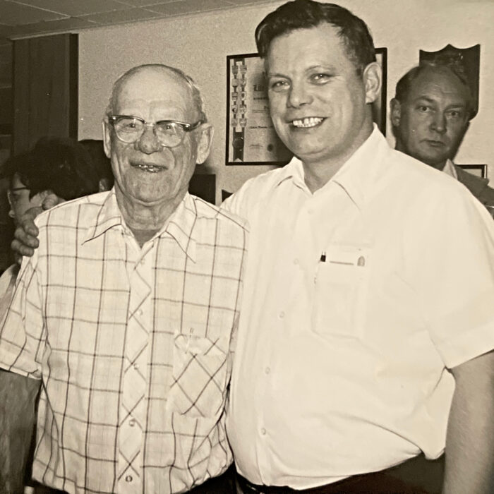 Black and white image of two men standing next to eachother. The younger man has his arm around the older man. Both men smile