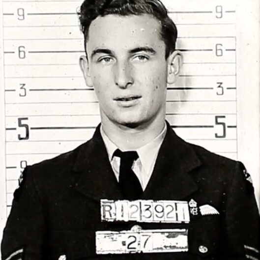 Charles Anthony Griffiths, c1940-1944, Canadian Virtual War Memorial Black and white image of Charles A. Griffiths standing in front of a measuring board. He holds his RCAF number sign in front of his chest. He wears a suit jacket, a button-up shirt, and a tie. His hair is short and styled