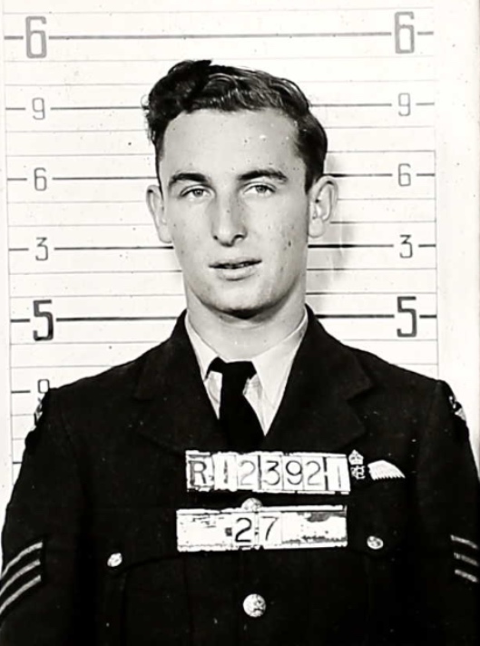 Black and white image of Charles A. Griffiths standing in front of a measuring board. He holds his RCAF number sign in front of his chest. He wears a suit jacket, a button-up shirt, and a tie. His hair is short and styled