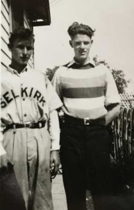 A black and white image of two young men standing looking at the camera. The young man on the right wears a wide-striped shirt and black pants. His hands are in his pockets. The young man on the left wears a baseball style button up shirt with the words "Selkirk" across the front.