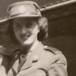 Black and white image of a woman in military uniform. She has short curly hair and wears a hat. She is looking at the camera and smiling