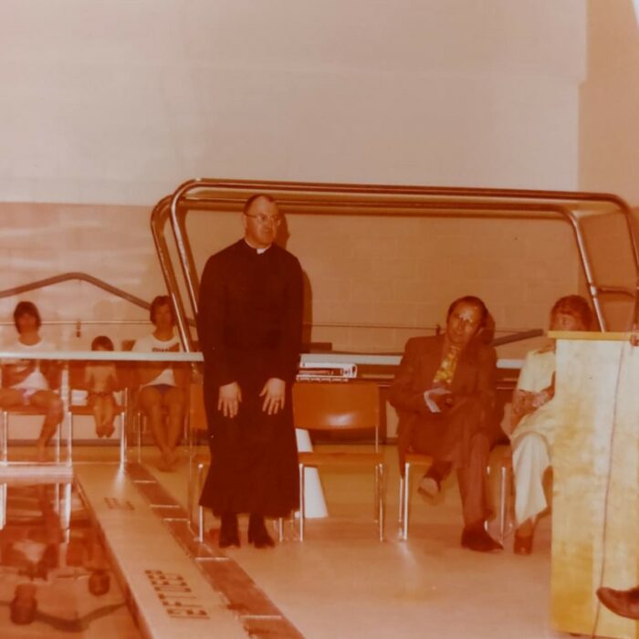 Colour image of a priest wearing cassocks standing next to an indoor pool. There are people in the background watching