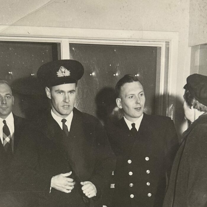 A black and white image of four people standing in front of a window. The three men are wearing suits and ties. One is wearing a military uniform. There is a woman on the right who is turned away from the camera.