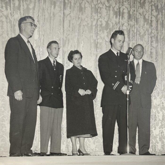 A black and white photograph of five people standing on a stage. There are two men on the left, a woman at the center, and two men on the right. One man stands at a microphone