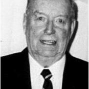 A black and white image of a man in a black suit. He wears a white shirt and a black tie. He looks a the camera