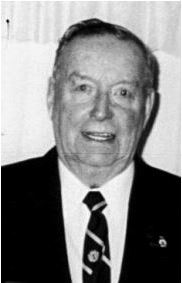 A black and white image of a man in a black suit. He wears a white shirt and a black tie. He looks a the camera
