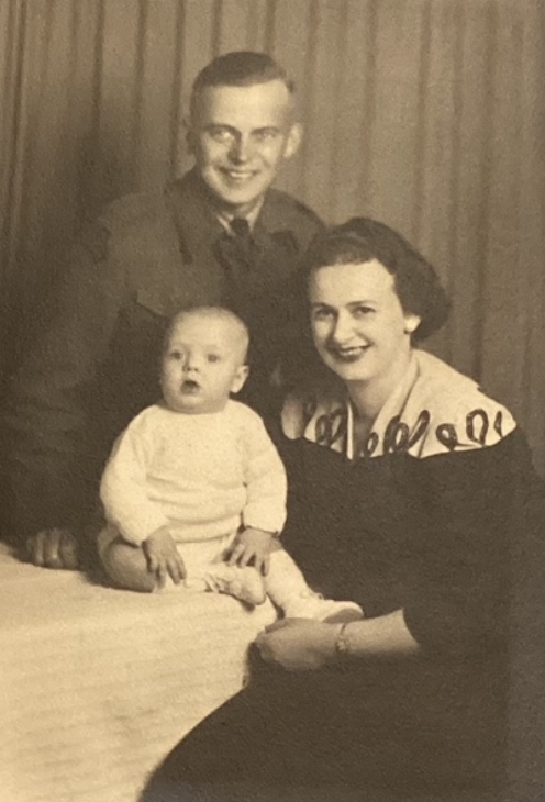 A black and white image of a young family. The mother holds her baby son in front of her and behind them both is the father. Both parents smile at the camera.