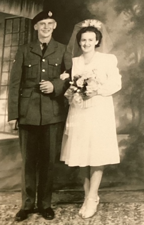 Black and white image of a bride and groom. The groom is wearing a military uniform and the bride is wearing a tea length white wedding dress. She holds flowers and the arm of her husband
