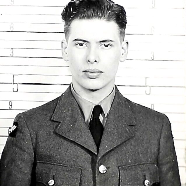 Jack Norquay R152593, 1943, Veterans Canada Black and white image of a young man dressed in military uniform. He is standing in front of a measuring chart to show his height. The young man looks at the camera