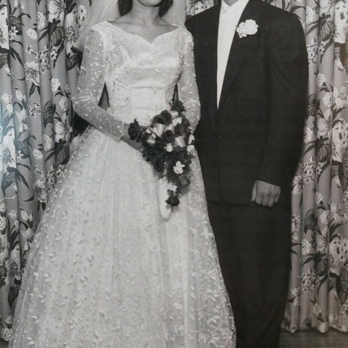 Black and white photo of a bride and groom on their wedding day. The bride wears a long white dress with a v neck and lace overlay. The sleeves are long and she holds a draping bouquet of flowers. The groom wears a white shirt with a dark suit jacket and pants. He has a white boutonniere on his jacket lapel