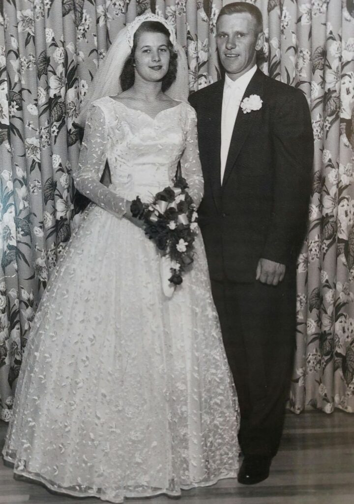 Black and white photo of a bride and groom on their wedding day. The bride wears a long white dress with a v neck and lace overlay. The sleeves are long and she holds a draping bouquet of flowers. The groom wears a white shirt with a dark suit jacket and pants. He has a white boutonniere on his jacket lapel