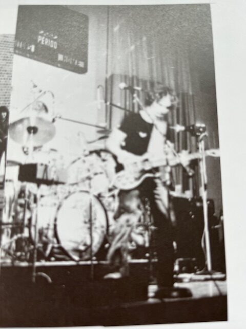 black and white photo of a singer singing on stage. He holds a guitar and there is a large drum set on the stage behind him.