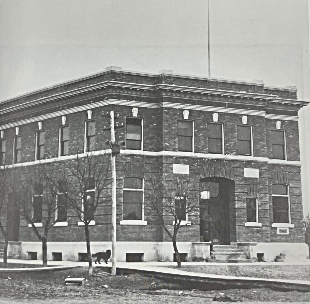 Black and white image of the post office from across main street. There is a dog walking down the sidewalk