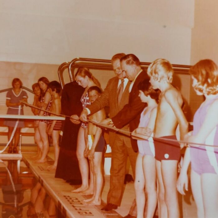 Sepia coloured image of people clustered around the edge of an indoor pool. The people hold a ribbon. Some of the people are children who wear swim suits, some are men who wear suits.