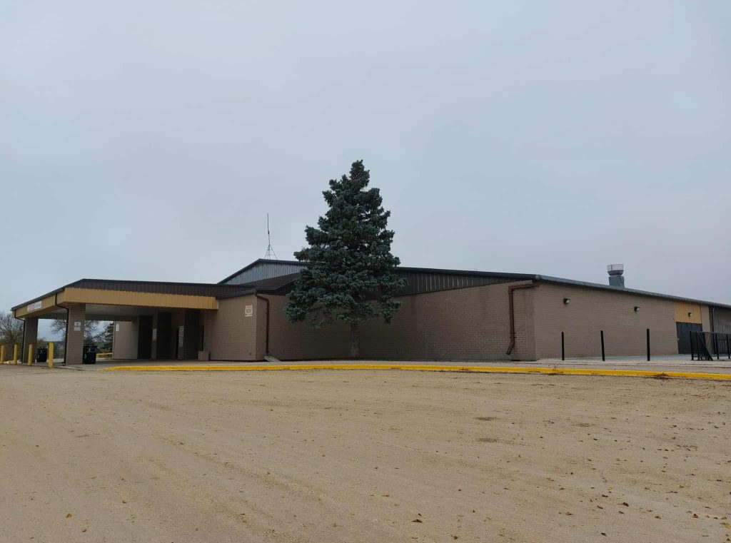 A colour image of the Selkirk Recreation Complex. The sky is overcast and there is a large evergreen tree in front of the building entrance