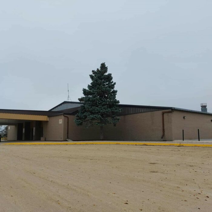 A colour image of the Selkirk Recreation Complex. The sky is overcast and there is a large evergreen tree in front of the building entrance