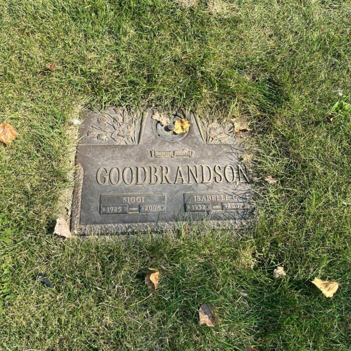 A colour image of a gravestone in the grass. The gravestone marker is made of metal and there are a few yellow leaves scattered on the grass